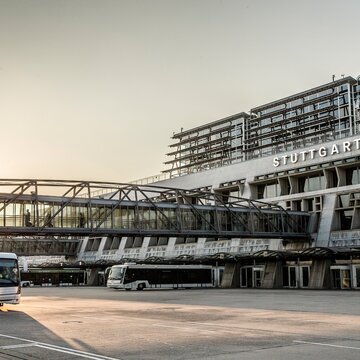 Außenansicht des Terminalgebäudes am Flughafen Stuttgart (Manfred Rommel Airport) mit Busverkehr im Vordergrund bei Sonnenaufgang.