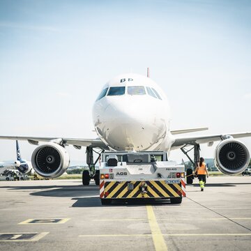Flugzeug wird von einem Schlepperfahrzeug auf dem Vorfeld des Flughafens Stuttgart gezogen, Mitarbeitende sichern den Vorgang.