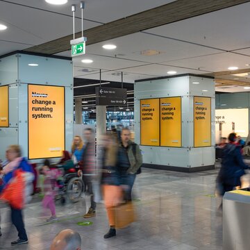 Menschen im Flughafen laufen an großen gelben Werbetafeln mit dem Text „Change a running system“vorbei. Das Wort "Never" auf den Plakaten ist durchgestrichen.