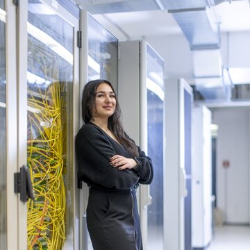 Eine junge Frau mit dunklen Haaren steht lächelnd vor einer Solaranlage auf dem Flughafengelände Stuttgart. Sie trägt eine dunkelblaue Jacke mit dem Logo „Stuttgart Airport“. Im Hintergrund hebt ein Flugzeug in den Himmel ab.