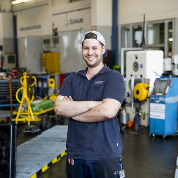 Lächelnder Mitarbeiter mit verschränkten Armen steht in einer Werkstatt des Flughafens Stuttgart.