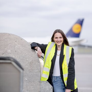 Junge Mitarbeiterin mit Warnweste lehnt entspannt an Flughafen-Ausrüstung, im Hintergrund steht ein Flugzeug.