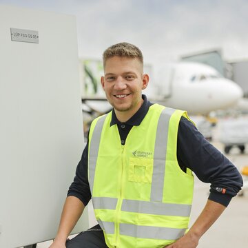 Ein junger Mann in dunkler Arbeitskleidung und neongelber Warnweste mit der Aufschrift „Stuttgart Airport“ steht draußen auf dem Flughafengelände. Er lächelt freundlich in die Kamera und lehnt mit einem Bein an einem Technikgehäuse. Im Hintergrund ist ein Flugzeug zu sehen.