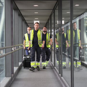 Drei Flughafenmitarbeiter in Warnwesten gehen mit Werkzeugkoffern durch eine gläserne Fluggastbrücke.