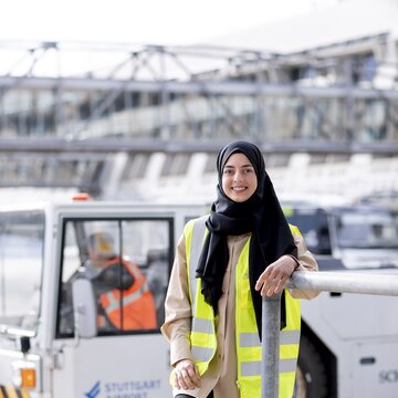Eine junge Frau mit schwarzem Kopftuch und gelber Warnweste steht lächelnd auf dem Vorfeld des Flughafens Stuttgart. Im Hintergrund ist ein Flughafenfahrzeug mit dem Logo „Stuttgart Airport“ zu sehen. Die Frau wirkt selbstbewusst und freundlich.