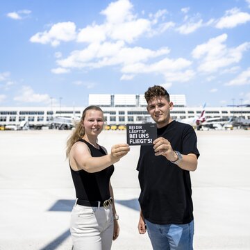 Zwei junge Personen halten ein Schild mit der Aufschrift „Bei dir läuft’s? Bei uns fliegt’s!“ vor dem Terminal des Flughafens Stuttgart.