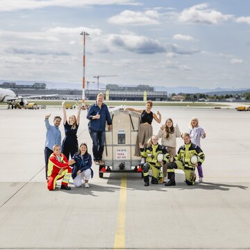 Mitarbeitende aus verschiedenen Bereichen posieren gut gelaunt gemeinsam auf dem Rollfeld vor einer Gepäckpalette.