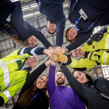 Gruppe lachender Flughafen-Mitarbeitender steht im Kreis und streckt die Hände gemeinsam in die Mitte.
