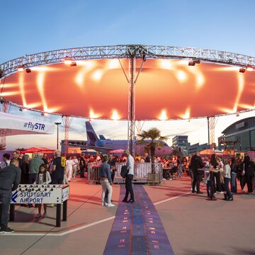 Menschenmenge bei einer Abendveranstaltung unter beleuchtetem Zeltdach auf dem Gelände des Flughafens Stuttgart mit Blick auf ein Flugzeug im Hintergrund.