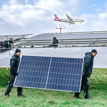  Mitarbeiter installieren Solarmodule am Flughafen Stuttgart, im Hintergrund fliegt ein Flugzeug über die Anlage.