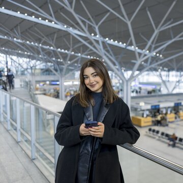 Junge Frau lehnt mit Smartphone an einem Geländer in der lichtdurchfluteten Abflughalle des Flughafens Stuttgart.