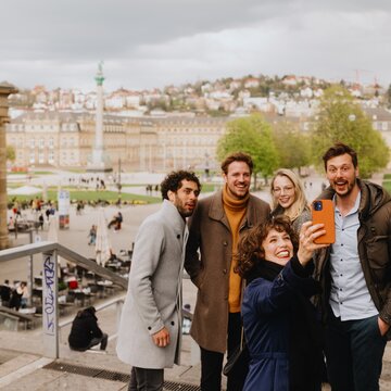 Eine Gruppe junger Menschen macht ein fröhliches Selfie auf der Treppe vor dem Schlossplatz in Stuttgart.