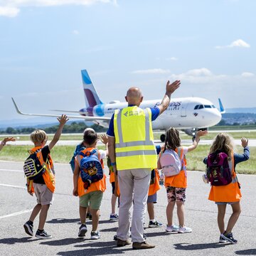 Kinder in orangefarbenen Sicherheitswesten winken einem Flugzeug auf dem Flughafen Stuttgart zu.